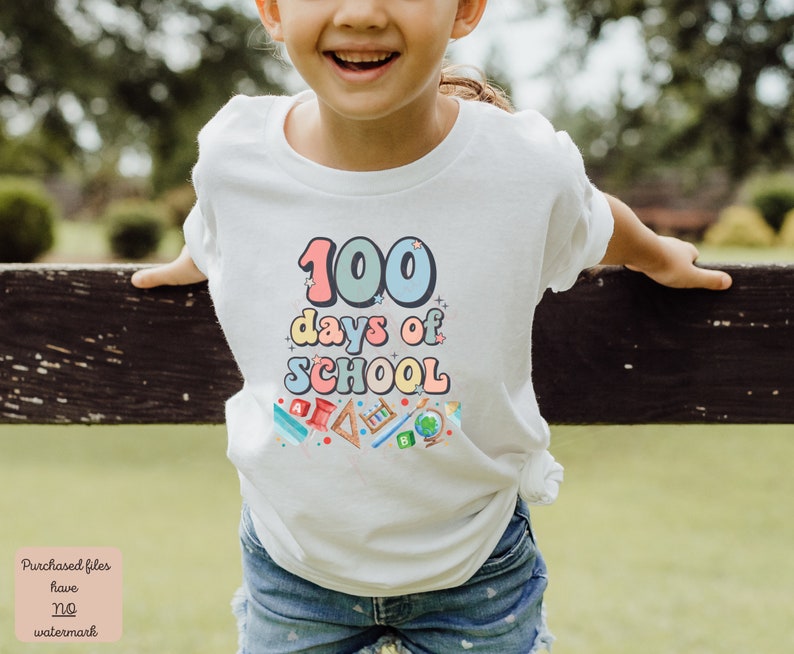 K&ouml;nnte beinhalten: Ein wei&szlig;es T-Shirt mit einer bunten Grafik, auf der "100 Tage Schule" steht, umgeben von Schulutensilien. Das Shirt wird von einem Kind getragen.