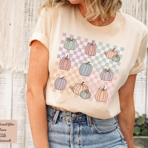 May include: A light beige t-shirt featuring a graphic design of pastel coloured pumpkins on a checkered background. The pumpkins are in various shades of pink, blue, and purple.