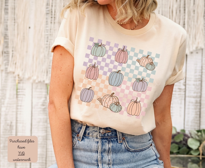 May include: A light beige t-shirt featuring a graphic design of pastel coloured pumpkins on a checkered background. The pumpkins are in various shades of pink, blue, and purple.