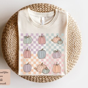 May include: A beige t-shirt with a checkered pattern featuring pastel-coloured pumpkins. The pumpkins are in various shades of pink, blue, and purple. The checkered pattern is in shades of light pink, light blue, and light purple.
