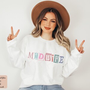 May include: A white sweatshirt with the word "MIDWIFE" printed in colorful, block letters. The letters are outlined in black and have a vintage, distressed look. The model is wearing a brown hat and smiling.