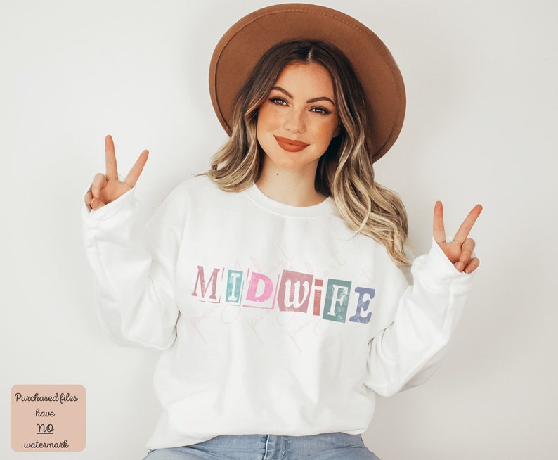 May include: A white sweatshirt with the word "MIDWIFE" printed in colorful, block letters. The letters are outlined in black and have a vintage, distressed look. The model is wearing a brown hat and smiling.