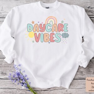 May include: White sweatshirt with the words "DAYCARE VIBES" in colorful letters. The design includes a rainbow, sun, stars, and clouds. The sweatshirt has long sleeves and a crew neck.