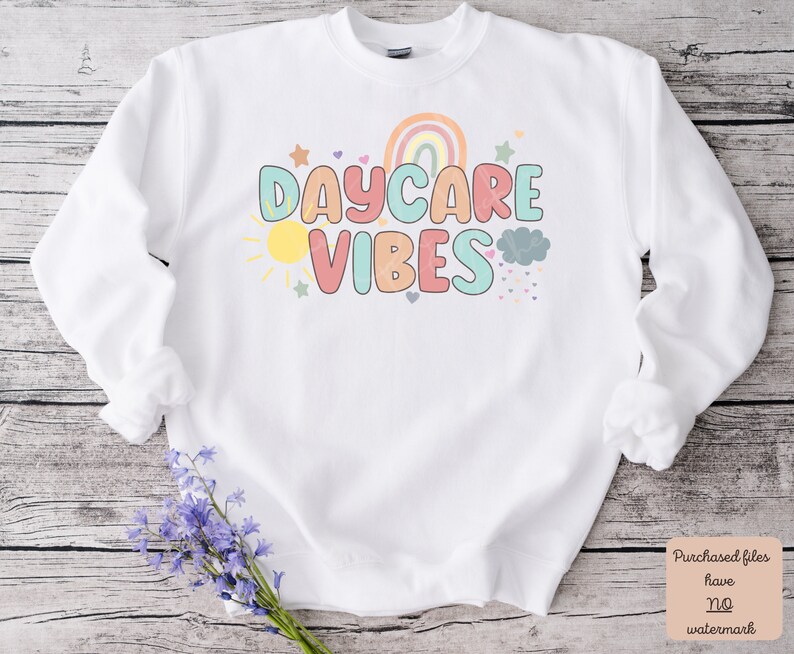 May include: White sweatshirt with the words "DAYCARE VIBES" in colorful letters. The design includes a rainbow, sun, stars, and clouds. The sweatshirt has long sleeves and a crew neck.