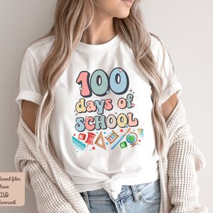 K&ouml;nnte beinhalten: Wei&szlig;es T-Shirt mit einer bunten Grafik, auf der "100 Tage Schule" steht, umgeben von Schulutensilien.