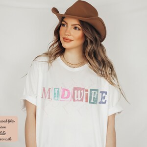 May include: A white t-shirt with the word "MIDWIFE" printed in colorful, block letters. The letters are made to look like magazine clippings.
