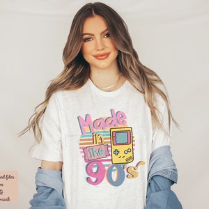 May include: White t-shirt with a graphic design that says "Made in the 90's" with a retro video game console illustration.