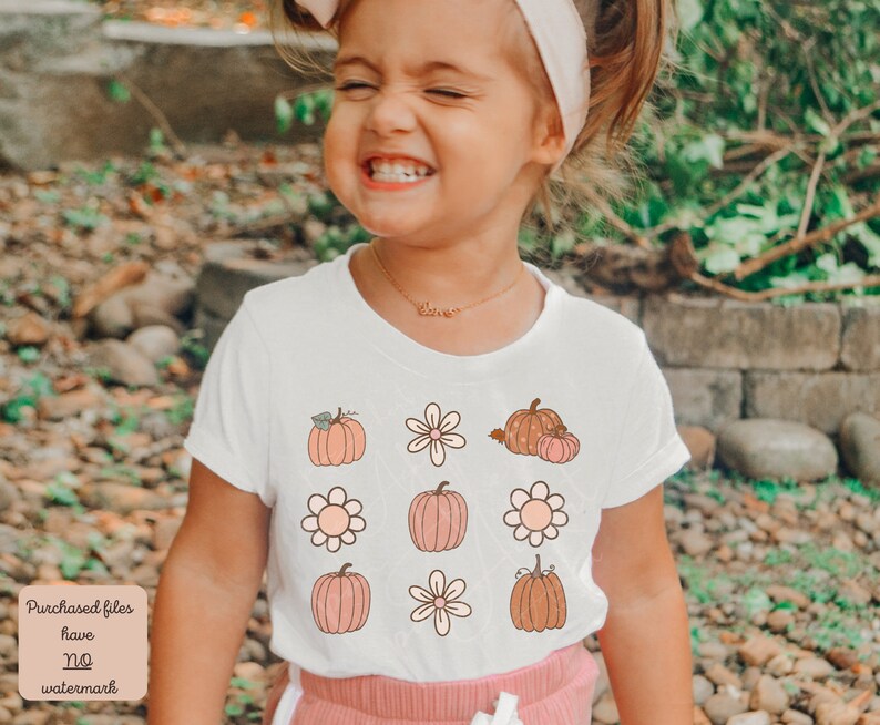Op de afbeelding: Een wit T-shirt met een schattig ontwerp met pompoenen, bloemen en bladeren in tinten oranje, bruin en roze. Het ontwerp wordt meerdere keren op het shirt herhaald.