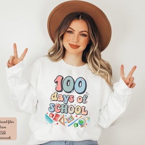 K&ouml;nnte beinhalten: Wei&szlig;er Pullover mit einer bunten Grafik, auf der "100 Tage Schule" steht, umgeben von Schulutensilien.