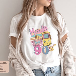 May include: White t-shirt with a graphic design that says "Made in the 90's" with a retro video game controller graphic.