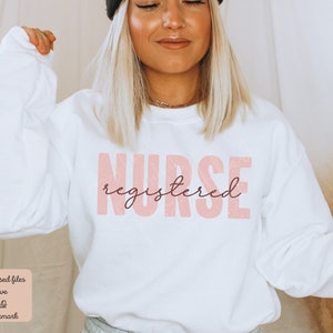 Puede incluir: Sudadera blanca con un gr&aacute;fico rosa que dice "NURSE registered" en una fuente desgastada.