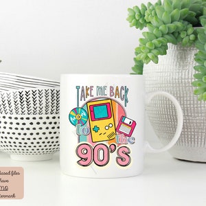 K&ouml;nnte beinhalten: Wei&szlig;e Keramiktasse mit einem bunten Grafikdesign, das eine Retro-Videospielkonsole, eine Diskette und eine CD zeigt. Der Text "Take Me Back to the 90's" ist auf die Tasse gedruckt.