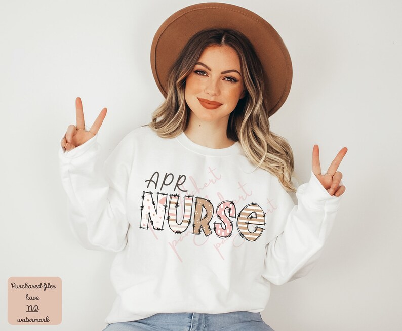 May include: White sweatshirt with the text "APR  NURSE" in pink and brown with a striped and polka dot pattern. The text is surrounded by a black outline.