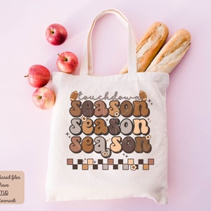 May include: A white tote bag with a brown, orange, and beige graphic that says "touchdown season" with a football and flowers. The bag is on a pink background with apples and bread.
