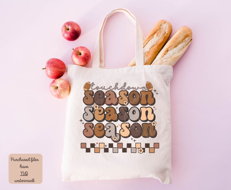 May include: A white tote bag with a brown, orange, and beige graphic that says "touchdown season" with a football and flowers. The bag is on a pink background with apples and bread.