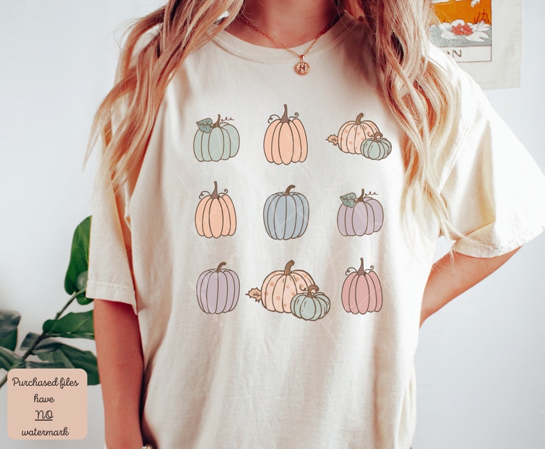 May include: A cream colored t-shirt with a pattern of nine pastel colored pumpkins. The pumpkins are in various shades of blue, pink, and orange.