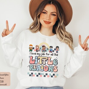 Pu&ograve; includere: Felpa bianca con un arcobaleno e una grafica di bambini dei cartoni animati. Il testo sulla felpa recita "Adoro il mio lavoro per tutte le piccole ragioni".