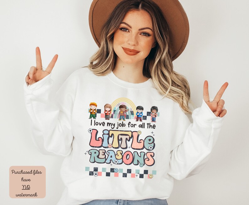 Pu&ograve; includere: Felpa bianca con un arcobaleno e una grafica di bambini dei cartoni animati. Il testo sulla felpa recita "Adoro il mio lavoro per tutte le piccole ragioni".