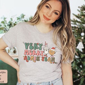 May include: A gray t-shirt with a Christmas-themed design featuring the text "Very Merry Admin Team" in a retro font. The design includes a cartoon milk carton and a cookie with a Santa hat.