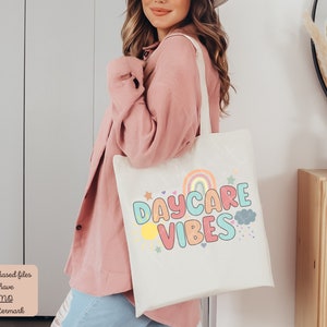 May include: Cream-colored tote bag with the words "DAYCARE VIBES" in colorful bubble letters, a rainbow, sun, stars, and clouds. The bag has a light-colored handle and is being held by a person wearing a pink shirt.