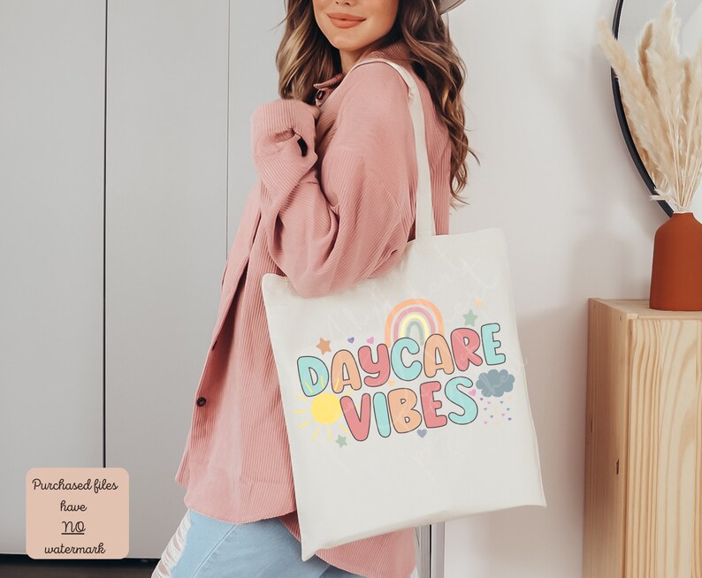 May include: Cream-colored tote bag with the words "DAYCARE VIBES" in colorful bubble letters, a rainbow, sun, stars, and clouds. The bag has a light-colored handle and is being held by a person wearing a pink shirt.