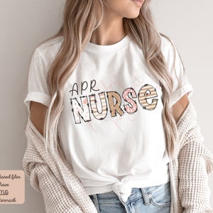 May include: White t-shirt with the text "APR NURSE" in brown and pink letters with a patterned design. The letters are outlined in black and have a stitched look.