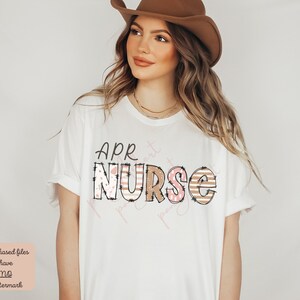 May include: A white t-shirt with the text "APR NURSE" in brown letters with pink and white patterned accents. The letters have a distressed look.