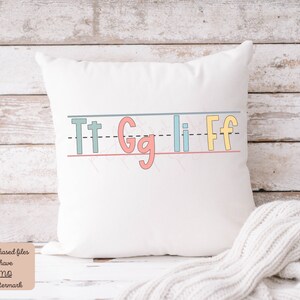 Pu&ograve; includere: Cuscino bianco con un designo alfabetico colorato. Le lettere sono "Tt Gg Li Ff" e sono scritte in un font da bambino. Le lettere sono su una linea con una linea tratteggiata sopra e una linea continua sotto.