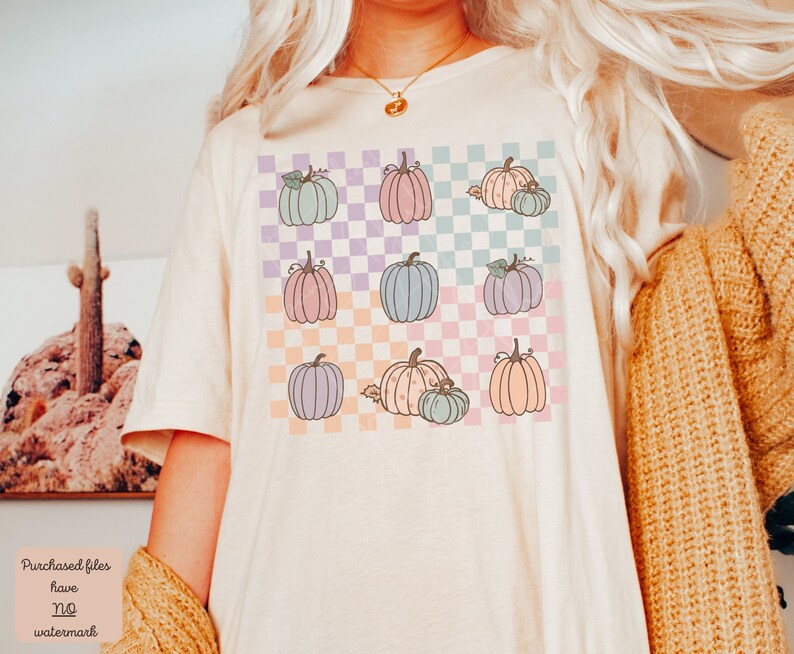 May include: A white t-shirt with a checkered pattern in pastel colours. The pattern features a repeated design of pastel pumpkins with stems and leaves.