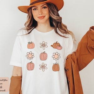Op de afbeelding: Een wit T-shirt met een grafisch ontwerp van pompoenen en bloemen in tinten oranje, roze en bruin. Het ontwerp wordt negen keer op het shirt herhaald.