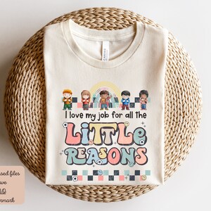 Pu&ograve; includere: Una maglietta beige con un design grafico colorato che presenta un gruppo di bambini e il testo "Amo il mio lavoro per tutte le PICCOLE RAGIONI".