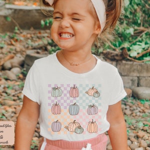 May include: A white t-shirt with a checkered pattern featuring pastel coloured pumpkins. The shirt has a crew neck and short sleeves.