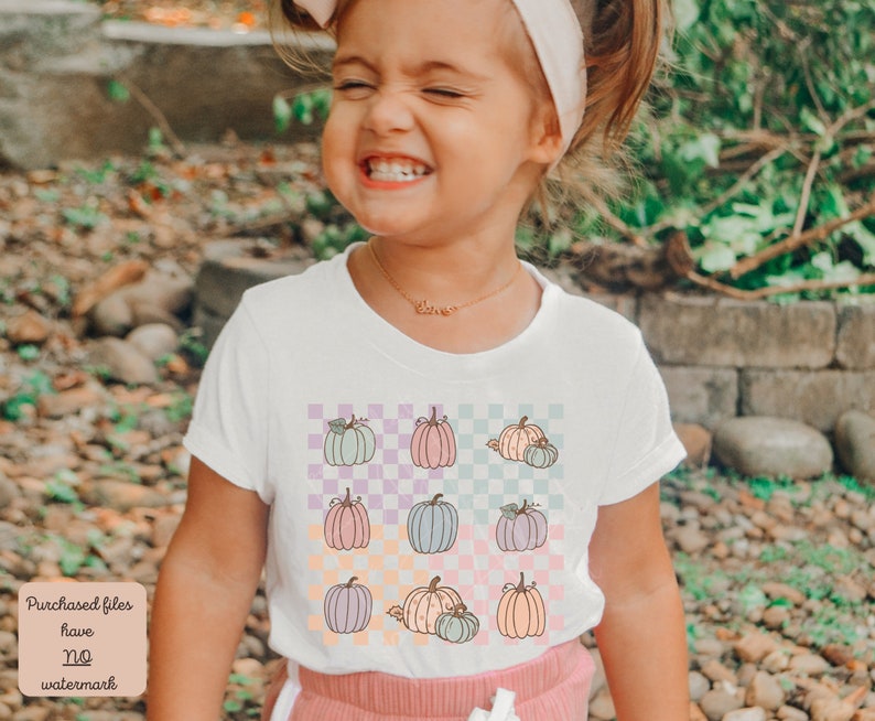 May include: A white t-shirt with a checkered pattern featuring pastel coloured pumpkins. The shirt has a crew neck and short sleeves.