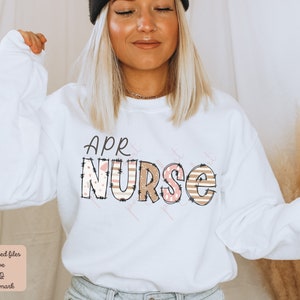 May include: White sweatshirt with the text "APR NURSE" in pink and brown letters with a patterned design. The letters are outlined in black.
