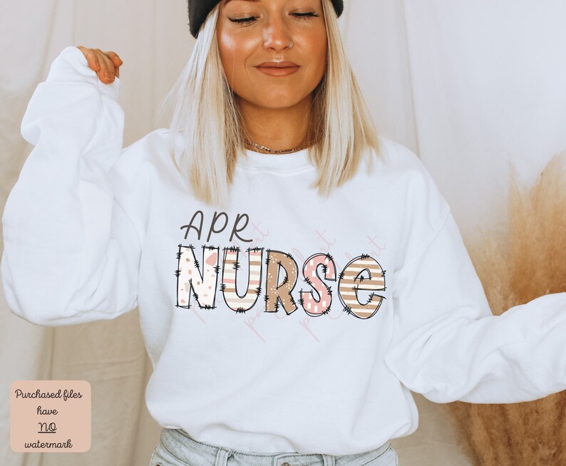 May include: White sweatshirt with the text "APR NURSE" in pink and brown letters with a patterned design. The letters are outlined in black.