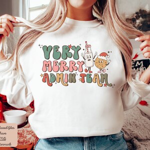 May include: White sweatshirt with a Christmas-themed design. The text reads "Very Merry Admin Team" in a retro font with a milk carton and cookie character.