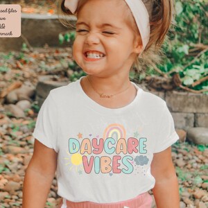 May include: A white t-shirt with the words "DAYCARE VIBES" in colorful bubble letters, with a rainbow, sun, stars, and cloud graphic. The shirt is worn by a young child.