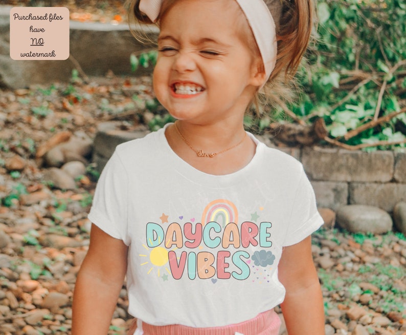 May include: A white t-shirt with the words "DAYCARE VIBES" in colorful bubble letters, with a rainbow, sun, stars, and cloud graphic. The shirt is worn by a young child.