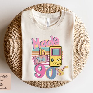 May include: A beige t-shirt with the text "Made in the 90's" in a retro font with a colorful sunset and a pixelated video game console graphic.