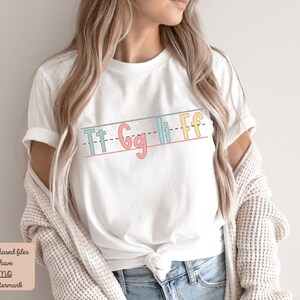 Pu&ograve; includere: Maglietta bianca con una grafica delle lettere "Tt Gg Li Ff" scritte in uno stile calligrafico colorato su uno sfondo a righe.