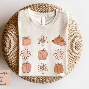 Op de afbeelding: Een beige T-shirt met een ontwerp van roze en oranje pompoenen en bloemen. Het ontwerp wordt herhaald in een rasterpatroon. Het T-shirt ligt op een geweven oppervlak.