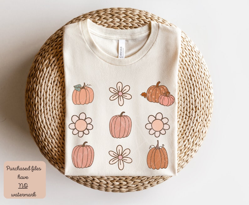 Op de afbeelding: Een beige T-shirt met een ontwerp van roze en oranje pompoenen en bloemen. Het ontwerp wordt herhaald in een rasterpatroon. Het T-shirt ligt op een geweven oppervlak.