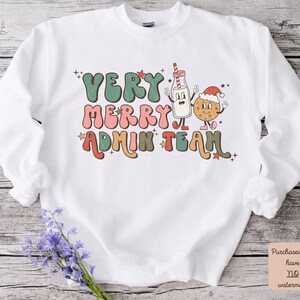May include: White sweatshirt with a retro-style graphic design featuring a milk carton and a cookie wearing Santa hats. The text reads "Very Merry Admin Team" in colorful, bubbly letters.