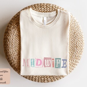May include: A beige t-shirt with the word "MIDWIFE" printed on it in colorful, distressed letters. The shirt is laid flat on a woven surface.