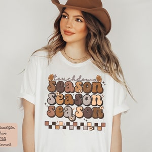 May include: White t-shirt with a brown, orange, and beige graphic that says "touchdown season season season". The graphic includes footballs and flowers.