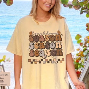 May include: A light yellow t-shirt with a retro-style graphic that says "Touchdown Season" in brown, tan, and gray letters. The graphic is surrounded by small white flowers and a checkered pattern.