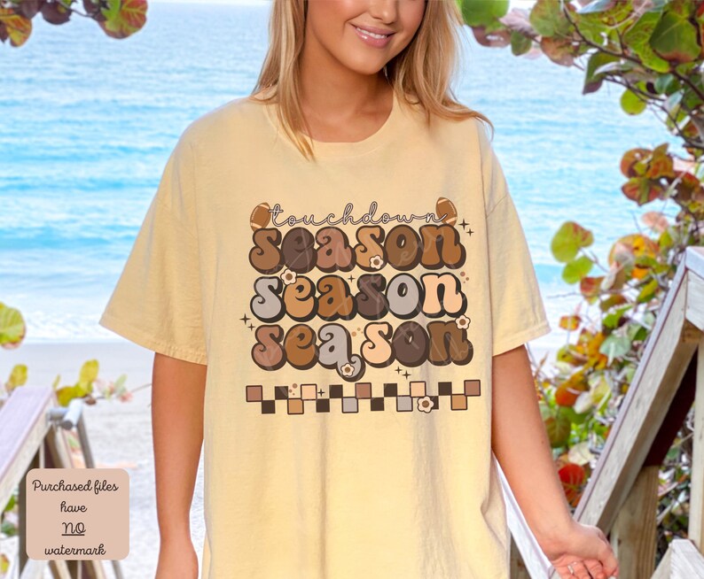 May include: A light yellow t-shirt with a retro-style graphic that says "Touchdown Season" in brown, tan, and gray letters. The graphic is surrounded by small white flowers and a checkered pattern.