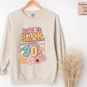 May include: A beige sweatshirt with the text "Take Me Back to the 90's" in colorful retro font. The text is surrounded by a colorful sunburst design and flowers.