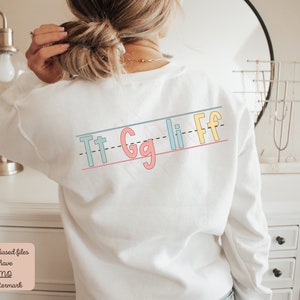 Pu&ograve; includere: Felpa bianca con una grafica colorata delle lettere "Tt Gg Ll Ff" stampate sul retro. Le lettere sono in uno stile di scrittura divertente e sono disposte in linea.