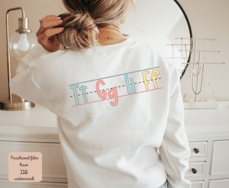 Pu&ograve; includere: Felpa bianca con una grafica colorata delle lettere "Tt Gg Ll Ff" stampate sul retro. Le lettere sono in uno stile di scrittura divertente e sono disposte in linea.